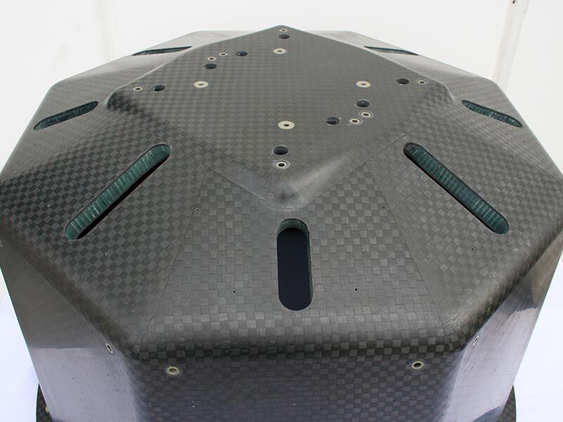 Closeup image of the top of the octagon section of the smallsat launch adapter structure. There are machined  holes, metallic inserts, and rectangular-shaped cutouts showing the core structure with potting material for a smooth finish inside the cutouts. The exterior is a carbon fiber weave with a matte finish. The background is white.